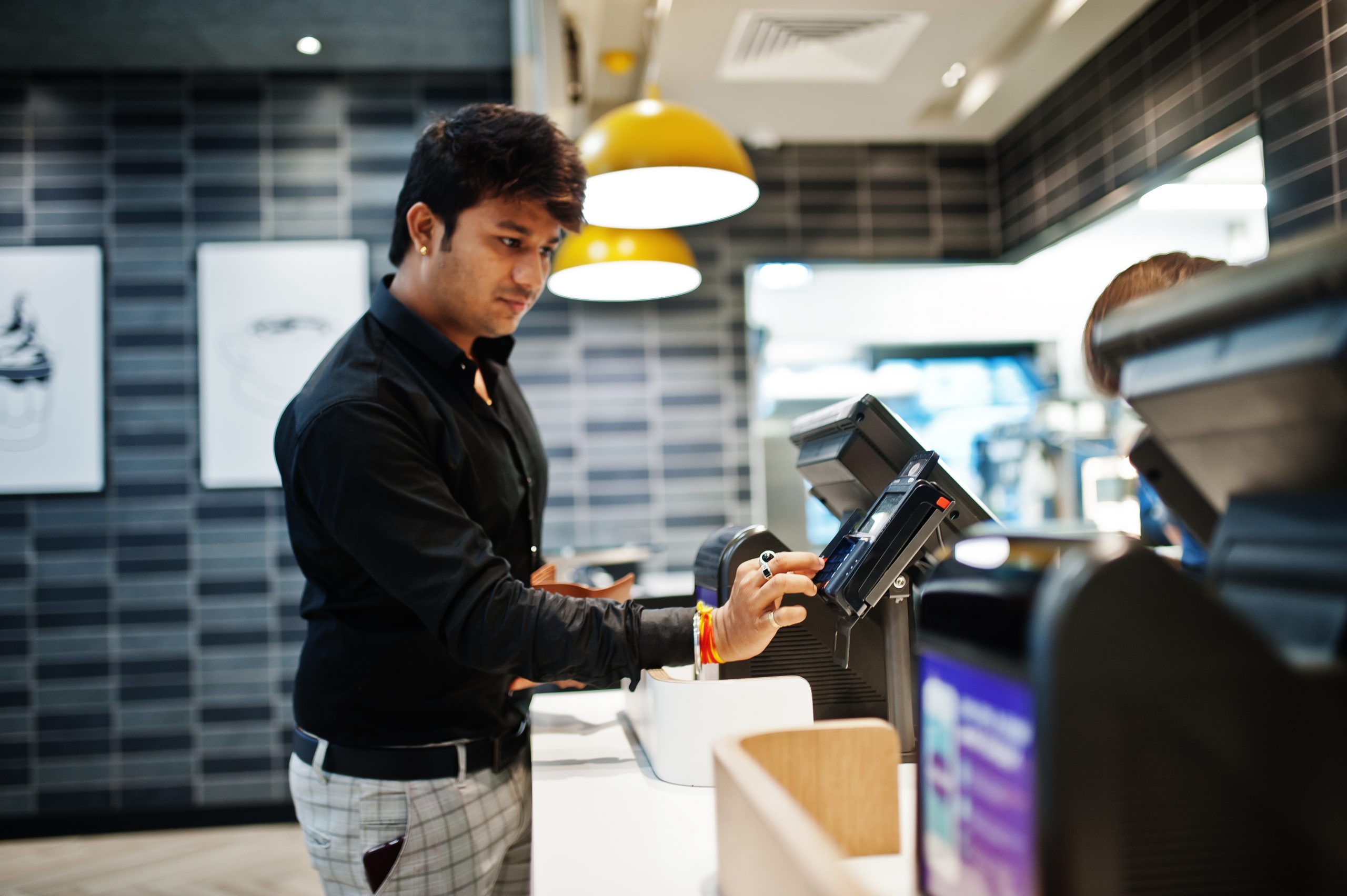 Man holding pay by credit card at cash desk with order screen and payment terminal in food cafe.